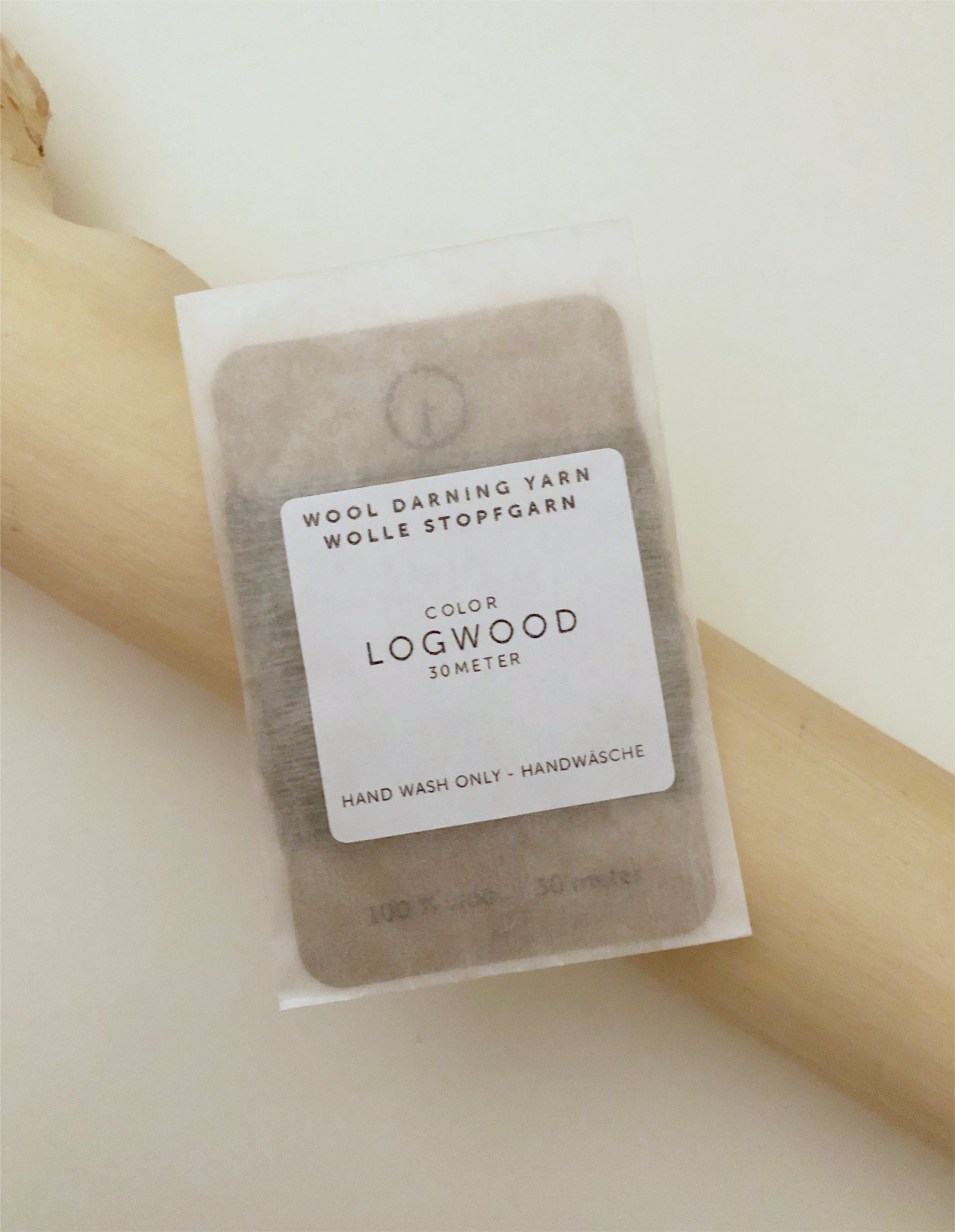 A 30-meter card of 100% wool darning yarn in logwood (dark beige/grey) colour, neatly wound and ready for mending knit garments. The yarn is packaged in a small, eco-friendly parchment paper bag.