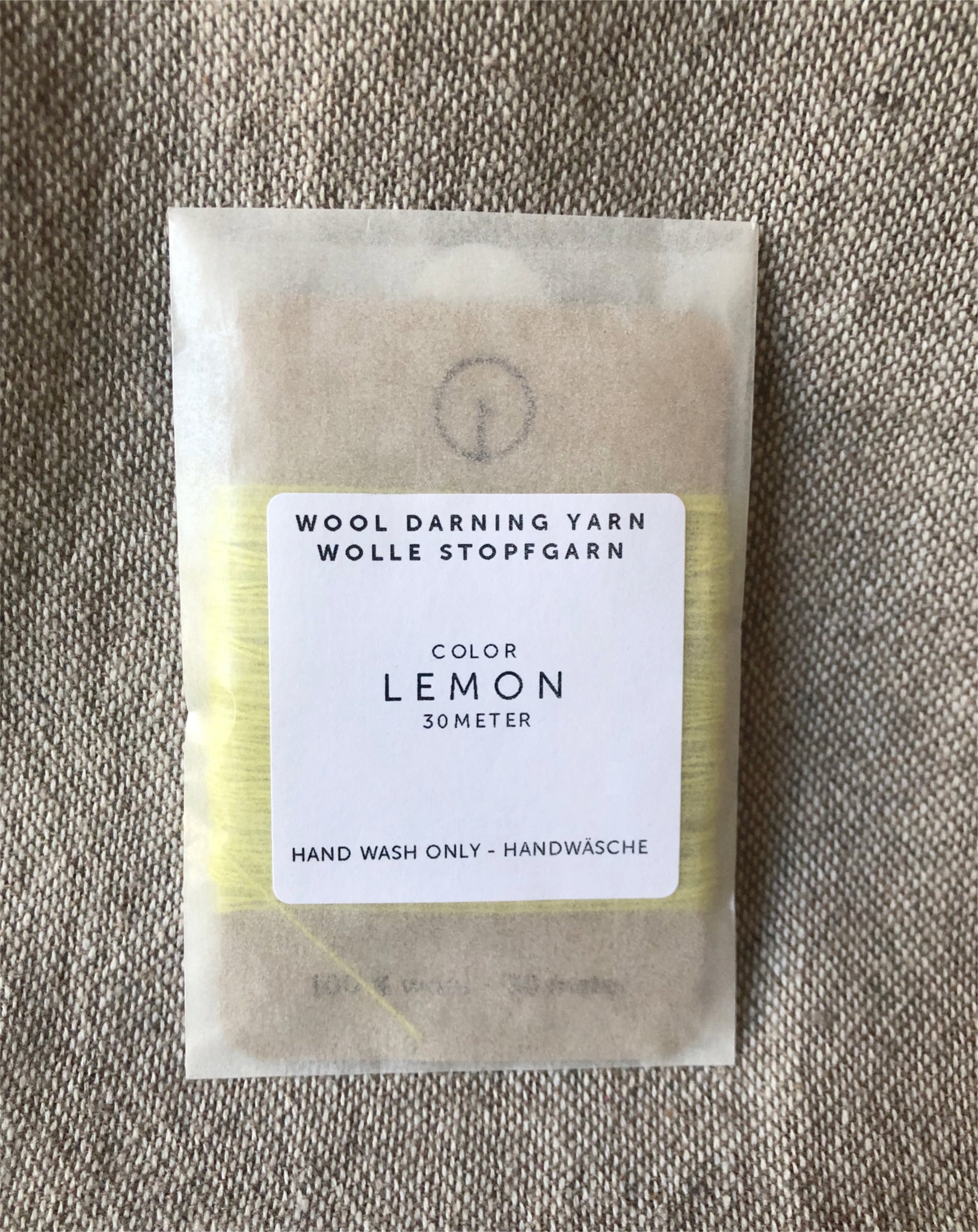 A 30-meter card of 100% wool darning yarn in light yellow lemon colour neatly wound and ready for mending knit garments. The yarn is packaged in a small, eco-friendly parchment paper bag.