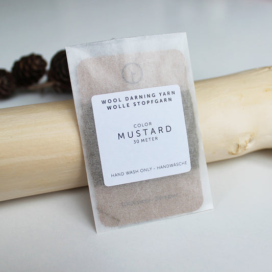 A 30-meter card of 100% wool darning yarn in mustard colour, neatly wound and ready for mending knit garments. The yarn is packaged in a small, eco-friendly parchment paper bag.