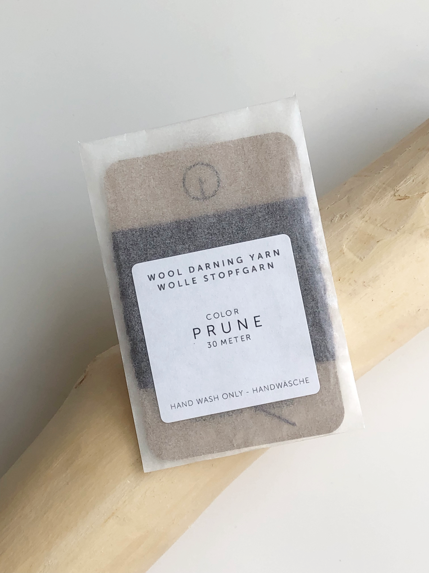 A 30-meter card of 100% wool darning yarn in prune purple colour, neatly wound and ready for mending knit garments. The yarn is packaged in a small, eco-friendly parchment paper bag.