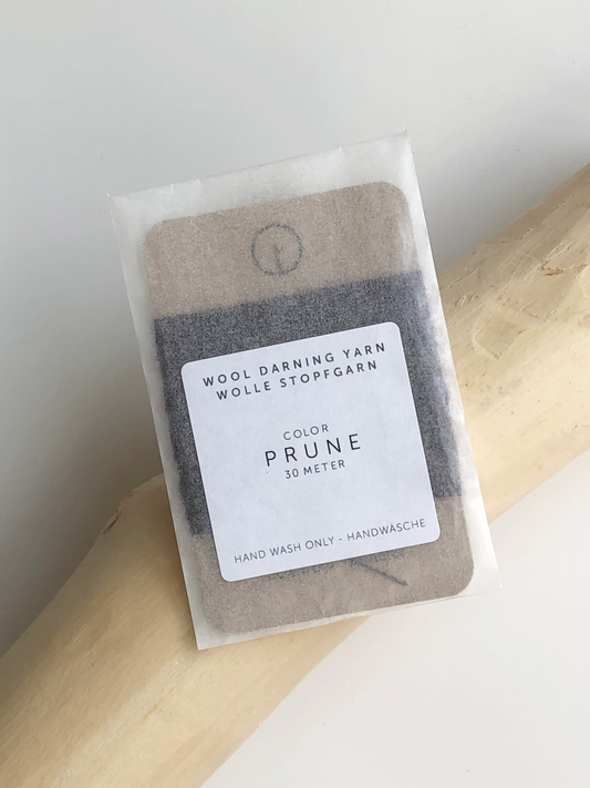 A 30-meter card of 100% wool darning yarn in prune purple colour, neatly wound and ready for mending knit garments. The yarn is packaged in a small, eco-friendly parchment paper bag.